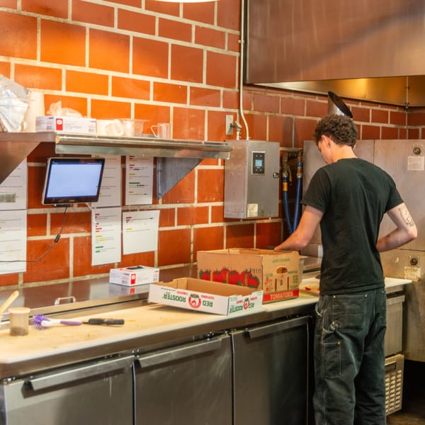 A chef in a black shirt and jeans unpacks tomatoes in a commercial kitchen with brick walls.