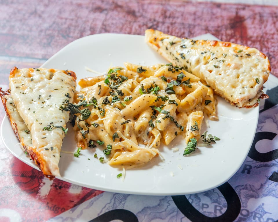 Creamy penne pasta with fresh basil and two slices of cheesy garlic bread on a white plate.
