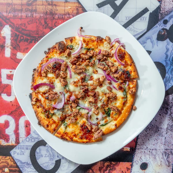 A round flatbread pizza with cheese, red onions, and pecans, on a white plate over a colorful map.
