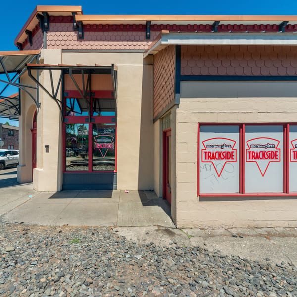 Exterior of Trackside Pizza restaurant with red-framed windows, a beige facade, and a gravel foreground.